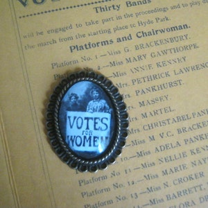 May include: A bronze-colored pin with a glass dome featuring a black and white image of two women and the text "Votes for Women".