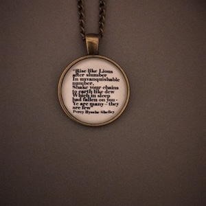 Könnte beinhalten: Eine bronzene Halskette mit einem runden Glasanhänger, auf dem ein Zitat von Percy Bysshe Shelley steht: "Erhebt euch wie Löwen nach dem Schlaf in unbezwingbarer Zahl, schüttelt eure Ketten, die wie Tau auf die Erde gefallen sind, als ihr schlieftet: Ihr seid viele, sie sind wenige".