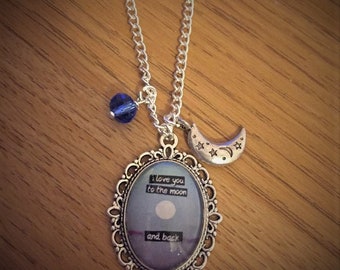 May include: A silver necklace with a charm that says "I love you to the moon and back". The charm is surrounded by a silver frame and has a blue background with a white moon. The necklace also has a small silver crescent moon charm and a blue crystal bead.