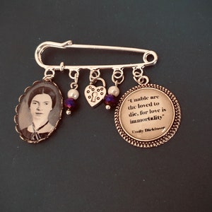 Puede incluir: Un broche de seguridad plateado con abalorios, incluyendo un medallón con una cerradura, un corazón y un abalorio redondo con una cita de Emily Dickinson: "Los amados no pueden morir, porque el amor es la inmortalidad."