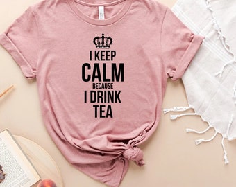 May include: A dusty rose t-shirt with rolled-up sleeves. The shirt features the text "I KEEP CALM BECAUSE I DRINK TEA" in black, topped with a crown graphic. The shirt is knotted at the bottom.
