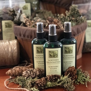 Puede incluir: Tres botellas de spray de vidrio con líquido verde y etiquetas que dicen "Northern Wildcrafters Northern Woods Potpourri Candles & Soy Melts Hand Picked & Packaged". Las botellas están sobre una superficie de madera con piñas y vegetación.