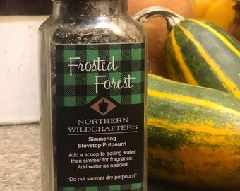 May include: A glass bottle with a black lid and a label reading "Frosted Forest" and "Northern Wildcrafters Simmering Stovetop Potpourri." The label features a green and black plaid design. Two green and yellow gourds are visible in the background.