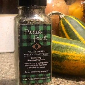 May include: A clear glass bottle with a black lid and a label that reads "Frosted Forest" and "Northern Wildcrafters Simmering Stovetop Potpourri." The label has a green and black plaid pattern. Two green and yellow gourds are in the background.