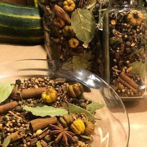 May include: A clear glass bowl and jars filled with a mix of spices, including cinnamon sticks, star anise, bay leaves, and small yellow gourds. The spices are arranged to create a visually appealing display, perfect for autumn decor or potpourri.