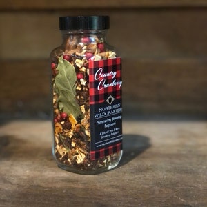 May include: A glass jar containing simmering stovetop potpourri, with a black lid and a label reading "Country Cranberry" on a red and black plaid background. The potpourri includes dried berries, leaves, and spices. The jar sits on a wooden surface.