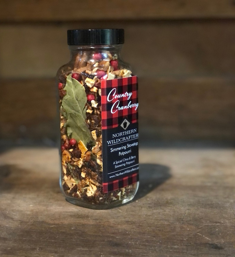 May include: A glass jar containing simmering stovetop potpourri, with a black lid and a label reading "Country Cranberry" on a red and black plaid background. The potpourri includes dried berries, leaves, and spices. The jar sits on a wooden surface.