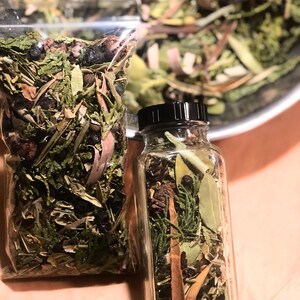 May include: A glass jar and a clear plastic bag containing a mixture of dried herbs, leaves, and berries. The jar has a black lid. The herbs are various shades of green and brown, with some dark berries.