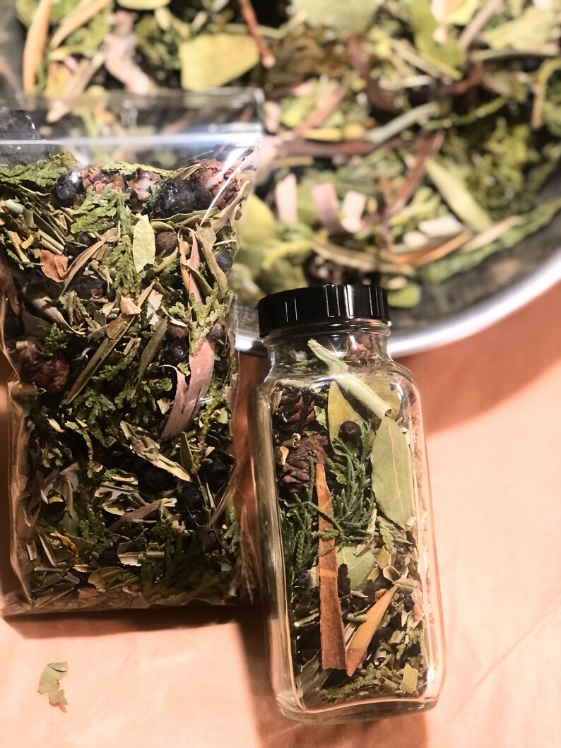 May include: A glass jar and a clear plastic bag containing a mixture of dried herbs, leaves, and berries. The jar has a black lid. The herbs are various shades of green and brown, with some dark berries.