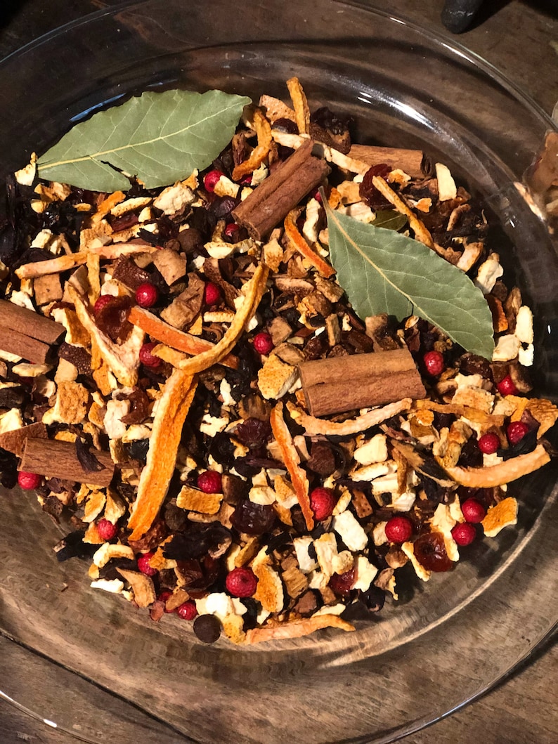 May include: A clear glass bowl containing a mixture of dried ingredients. The mix includes cinnamon sticks, orange peel, bay leaves, and red peppercorns. The ingredients are arranged in a visually appealing way.