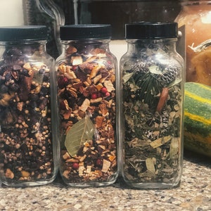 May include: Three glass jars filled with dried herbs and spices. The jars have black lids and contain a variety of ingredients, including berries, leaves, and cinnamon sticks. The jars are arranged on a countertop.