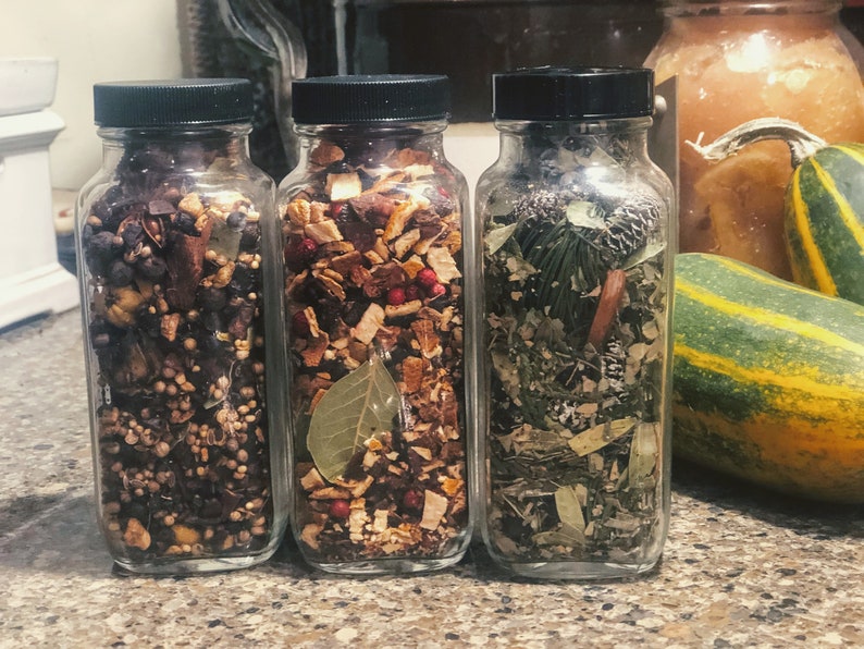 May include: Three glass jars filled with dried herbs and spices. The jars have black lids and contain a variety of ingredients, including berries, leaves, and cinnamon sticks. The jars are arranged on a countertop.