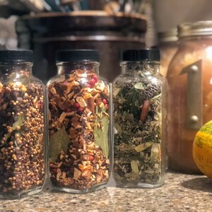 May include: Three glass jars filled with dried herbs and spices, each with a black lid. The jars contain a variety of ingredients, including small seeds, red berries, and green leaves. A decorative gourd and a large jar are in the background.