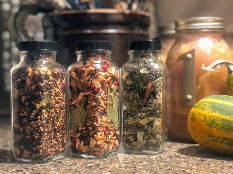 May include: Three glass jars filled with dried herbs and spices, each with a black lid. The jars contain a variety of ingredients, including small seeds, red berries, and green leaves. A decorative gourd and a large jar are in the background.