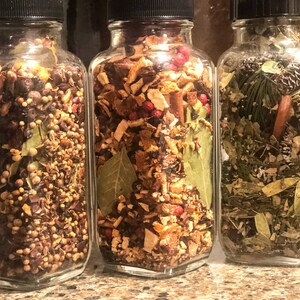 May include: Three glass jars with black lids, filled with a variety of dried herbs and spices. The jars contain a mix of brown, green, and red ingredients. The jars are displayed side by side, highlighting the different blends of ingredients.