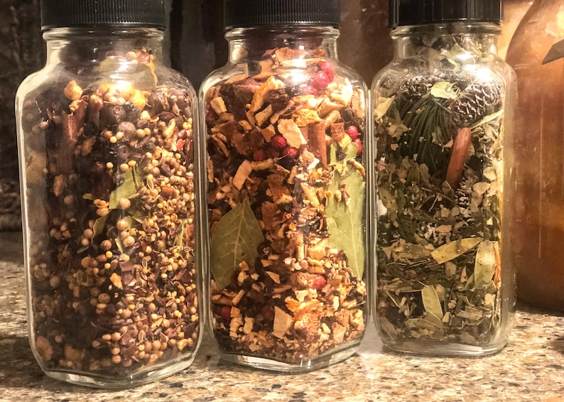 May include: Three glass jars with black lids, filled with a variety of dried herbs and spices. The jars contain a mix of brown, green, and red ingredients. The jars are displayed side by side, highlighting the different blends of ingredients.