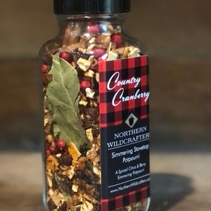 May include: A clear glass jar filled with a simmering stovetop potpourri. The jar has a black lid and a label with the text "Country Cranberry" and "Northern Wildcrafter". The potpourri includes dried citrus, berries, and a bay leaf.