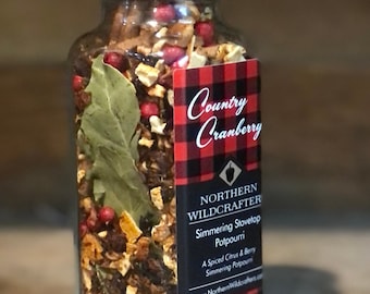 May include: A glass jar filled with simmering stovetop potpourri. The jar has a black lid and a label with the text "Country Cranberry" and "Northern Wildcrafter". The potpourri includes dried citrus, berries, and a bay leaf.