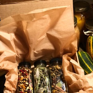 May include: Three glass bottles filled with dried herbs and spices, each with a black lid, are presented in a brown paper-lined box. The bottles contain a variety of colourful ingredients, including red berries, green leaves, and brown spices. The box is open, revealing the contents.