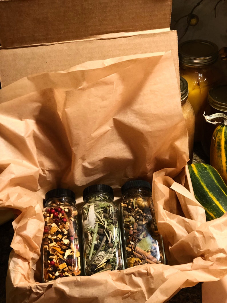 May include: Three glass bottles filled with dried herbs and spices, each with a black lid, are presented in a brown paper-lined box. The bottles contain a variety of colourful ingredients, including red berries, green leaves, and brown spices. The box is open, revealing the contents.