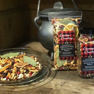 May include: A clear glass dish and two containers filled with a simmering stovetop potpourri. The potpourri includes dried orange slices, cinnamon sticks, bay leaves, and red berries. The containers have labels that read "Country Cranberry" and "Northern Wildcrafters".