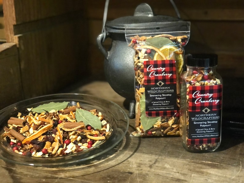 May include: A clear glass dish and two containers filled with a simmering stovetop potpourri. The potpourri includes dried orange slices, cinnamon sticks, bay leaves, and red berries. The containers have labels that read "Country Cranberry" and "Northern Wildcrafters".