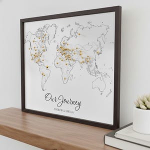 May include: Framed world map with gold push pins marking locations. The map is black and white with the words "Our Journey" and the names "Andrew & Amelia" in script. The frame is dark brown and the map is displayed on a wooden shelf.