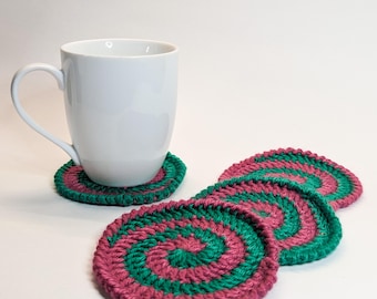 May include: Four crocheted coasters in pink and green. The coasters are round and have a spiral pattern. A white mug is sitting on one of the coasters.
