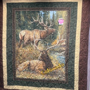 May include: A quilted wall hanging featuring a detailed image of two elk in a forest setting. The central image is framed by a green border, a tan border, and a brown outer border. The word "Colorado" is printed in gold on the image.