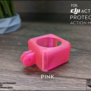 May include: A pink 3D printed protector for a DJI Action 2 camera. The protector is designed to mount the camera to an action mount. The text "FOR: DJI ACTION 2 PROTECTOR ACTION MOUNT" is printed on the image.