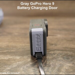 May include: Gray GoPro Hero 9 battery charging door replacement. The door is made of gray plastic and has the GoPro logo printed on it. The door is connected to a black charging cable.