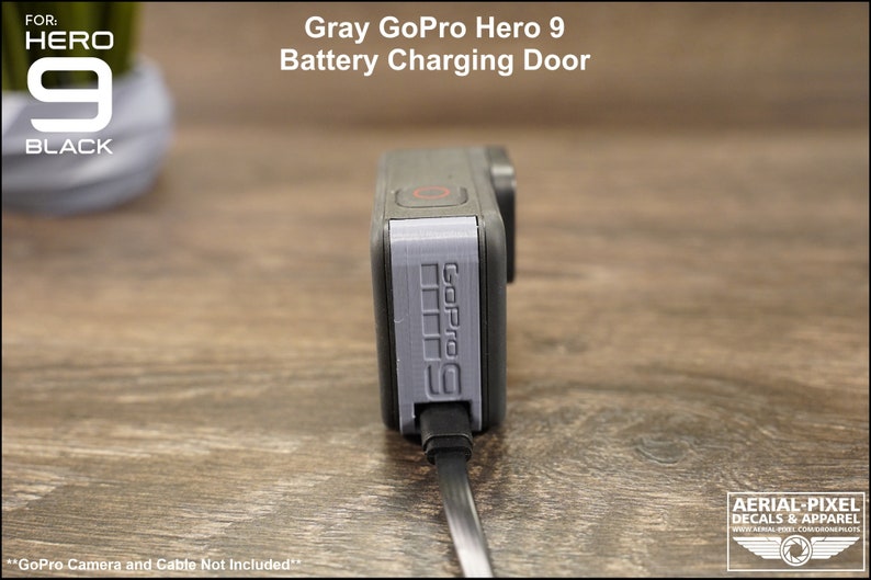 May include: Gray GoPro Hero 9 battery charging door replacement. The door is made of gray plastic and has the GoPro logo printed on it. The door is connected to a black charging cable.