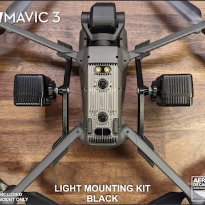 K&ouml;nnte beinhalten: Ein schwarzes Lichtmontage-Kit f&uuml;r eine DJI Mavic 3 Drohne. Das Kit enth&auml;lt zwei schwarze Lichthalterungen, die an den Armen der Drohne befestigt sind. Der Text "LIGHT MOUNTING KIT BLACK" ist sichtbar. Die Drohne ist grau mit schwarzen Propellern.
