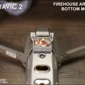 May include: A 3D printed mount for a DJI Mavic 2 drone, designed to hold a Firehouse Arc or Arc II strobe light. The mount is gray and is attached to the bottom of the drone. The strobe light is not included.
