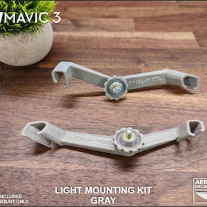 K&ouml;nnte beinhalten: Zwei graue Lichtmontages&auml;tze f&uuml;r DJI Mavic 3 Drohnen. Die S&auml;tze sind 3D-gedruckt und verf&uuml;gen &uuml;ber einen zentralen Schraubmechanismus und den Text "AERIAL-PIXEL". Das Bild enth&auml;lt auch den Text "LIGHT MOUNTING KIT GRAY".