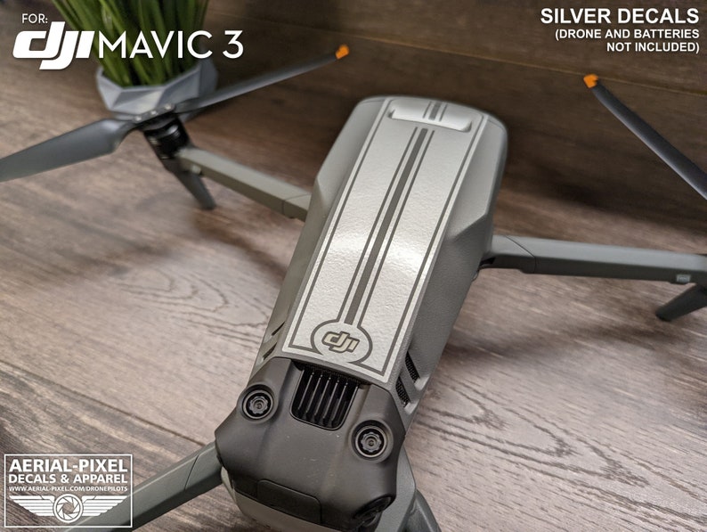 May include: Silver decals for a DJI Mavic 3 drone. The decals are two thin silver stripes running down the top of the drone. The drone is gray and black. The text "FOR: DJI MAVIC 3" is at the top of the image. The text "SILVER DECALS (DRONE AND BATTERIES NOT INCLUDED)" is at the top right of the image.