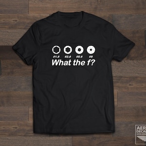Könnte beinhalten: Schwarzes T-Shirt mit einer Grafik von vier Kamera-Blendenstufen, beschriftet mit f/1.8, f/2.8, f/5.6 und f/8. Der Text "What the f?" ist unter der Grafik gedruckt.