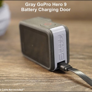 May include: Gray GoPro Hero 9 battery charging door. The door is attached to a gray GoPro Hero 9 camera. A black charging cable is plugged into the door. The text "GoPro Camera and Cable Not Included" is printed below the image.