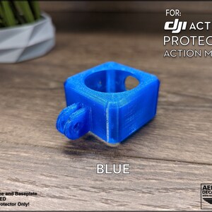 May include: Blue 3D printed protector for a DJI Action 2 camera. The protector is a square shape with a round opening for the camera lens. It has a GoPro-style mount on the bottom.  FOR: DJI ACTION 2 PROTECTOR ACTION MOUNT