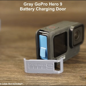 May include: Gray GoPro Hero 9 battery charging door replacement. The door is made of gray plastic and has the GoPro logo and the number 9 printed on it. The door is shown next to a black GoPro Hero 9 camera.