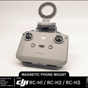 Op de afbeelding: Een grijze magnetische telefoonhouder voor DJI RC-N1, RC-N2 en RC-N3 afstandsbedieningen. De houder heeft een cirkelvormige telefoonhouder en is ontworpen om aan de afstandsbediening te bevestigen. De tekst "Magnetic Phone Mount" is zichtbaar.