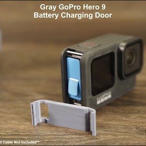 May include: Gray replacement battery charging door for a GoPro Hero 9 Black action camera. The door is made of plastic and has a small blue latch. The text "GoPro Camera and Cable Not Included" is visible at the bottom of the image.