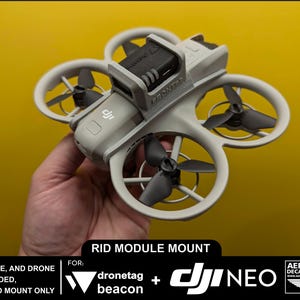 May include: A 3D printed mount for a dronetag beacon and a DJI Neo drone. The mount is white and has a black dronetag beacon attached to it. The text "RID MODULE MOUNT" is printed on the mount. The text "FOR: dronetag beacon + DJI NEO" is printed below the mount.