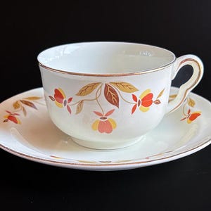 May include: White teacup and saucer set with a gold rim and autumn leaf design in orange, yellow, and brown. The cup has a curved handle and sits on a matching saucer. The set is on a black surface.