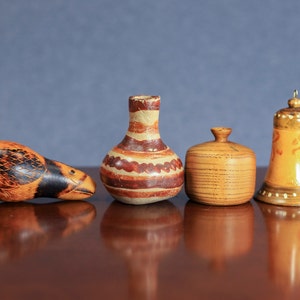 May include: Four decorative wooden objects on a dark brown surface. The objects include a bird-shaped carving, a small vase with brown and white stripes, a round wooden container, and a bell-shaped ornament with gold accents.