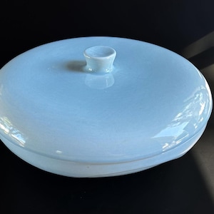 May include: A light blue ceramic lidded dish. The round dish has a matching lid with a small, rounded handle. The glossy surface reflects light, and the overall design is simple and elegant. The dish is likely for storage or display.