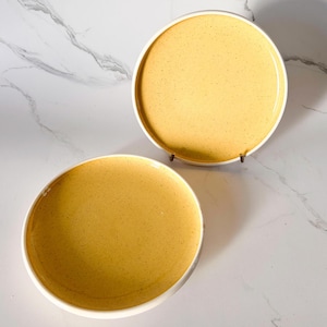 May include: Two round ceramic plates with a speckled yellow surface and a white rim. One plate is propped up on a metal stand. The plates are on a white marble surface.