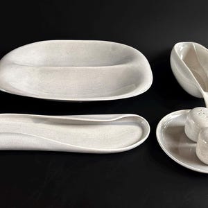 May include: A set of four speckled white ceramic serving dishes. Includes a long, curved tray, a divided oval dish, a gravy boat, and a small plate with two salt shakers. The pieces have a minimalist, modern design.