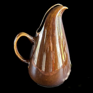 May include: A brown ceramic pitcher with a glossy finish. The pitcher has a curved handle and a spout designed for pouring. The body of the pitcher is a teardrop shape. The pitcher is likely for serving beverages.