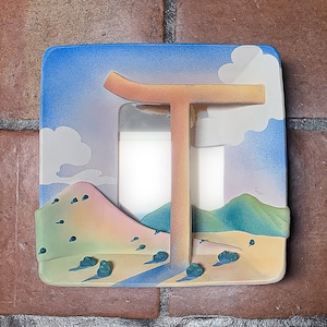 May include: A square, decorative light switch cover featuring a desert landscape design. The cover has a blue sky with clouds, a tan arch, and a colorful desert scene with mountains and bushes. The cover is mounted on a brick wall.